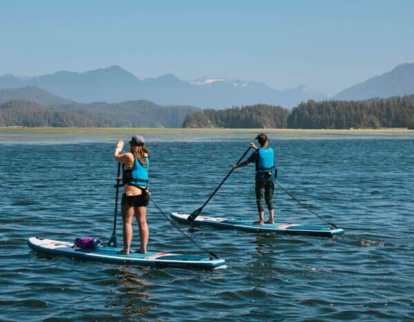 Planche de paddle gonflable Opti en eau avec deux personnes en paddle, paysage montagneux en fond.